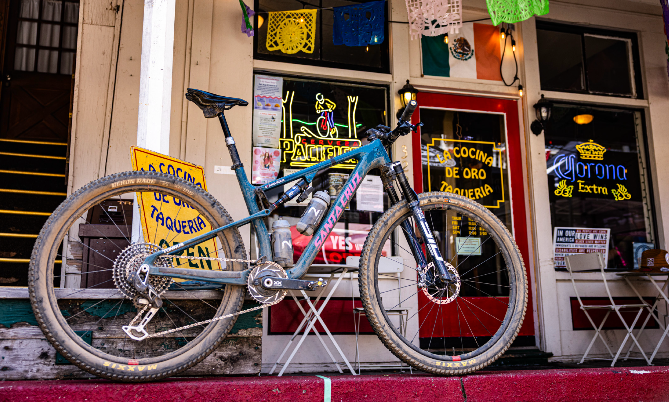 A Santa Cruz mountain bike, covered in trail dust, leans against a vibrant taqueria with colorful signage and decorations