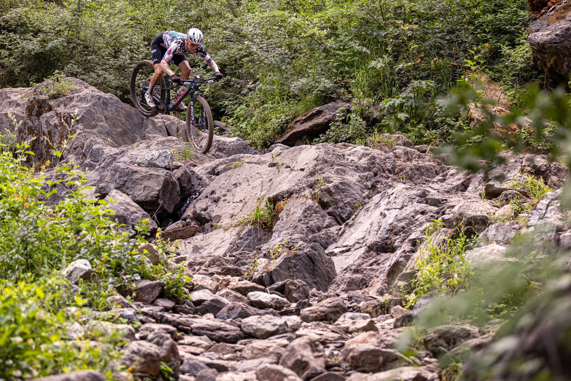 A mountain biker navigates a challenging rocky descent surrounded by lush greenery in an outdoor trail setting