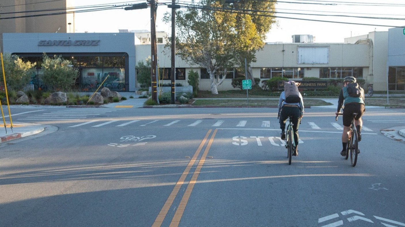 Two cyclists on a road approaching a Santa Cruz storefront, with marked bike lanes and a sunny backdrop