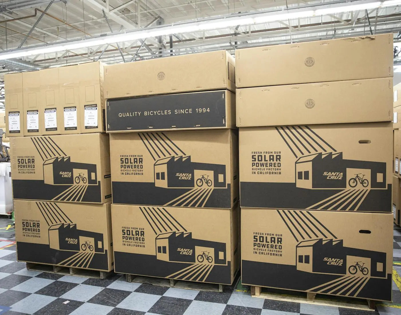 Stacks of Santa Cruz bicycle boxes labeled "Fresh from our solar-powered bicycle factory in California" and "Quality bicycles since 1994" in a warehouse setting