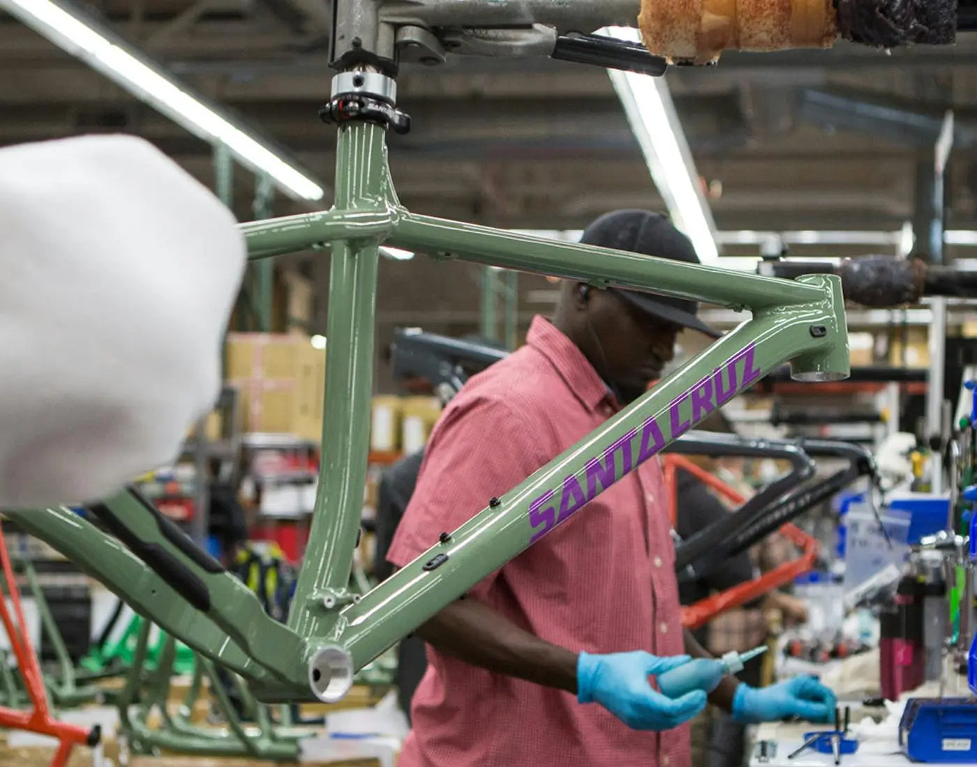 A worker wearing gloves assembles a green Santa Cruz bike frame in a busy workshop, surrounded by tools and other frames