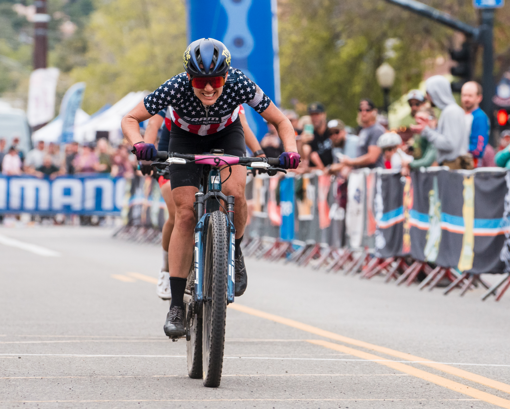 A cyclist wearing a USA jersey and helmet rides hard toward the finish line in a road race, cheered by a crowd.