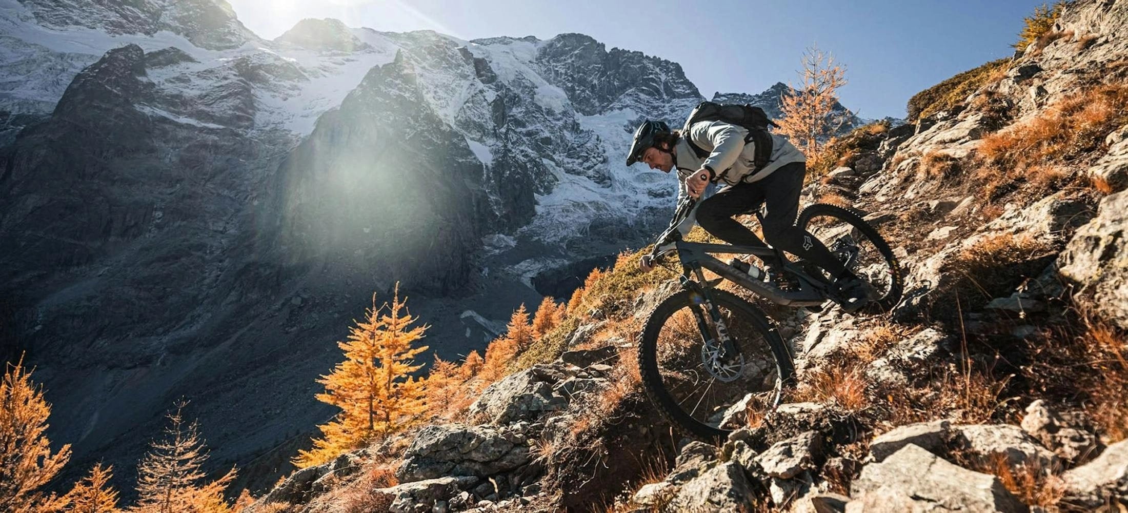 A mountain biker navigates a steep, rocky trail surrounded by golden autumn foliage, with a stunning backdrop of towering, snow-capped peaks illuminated by the sun's warm glow