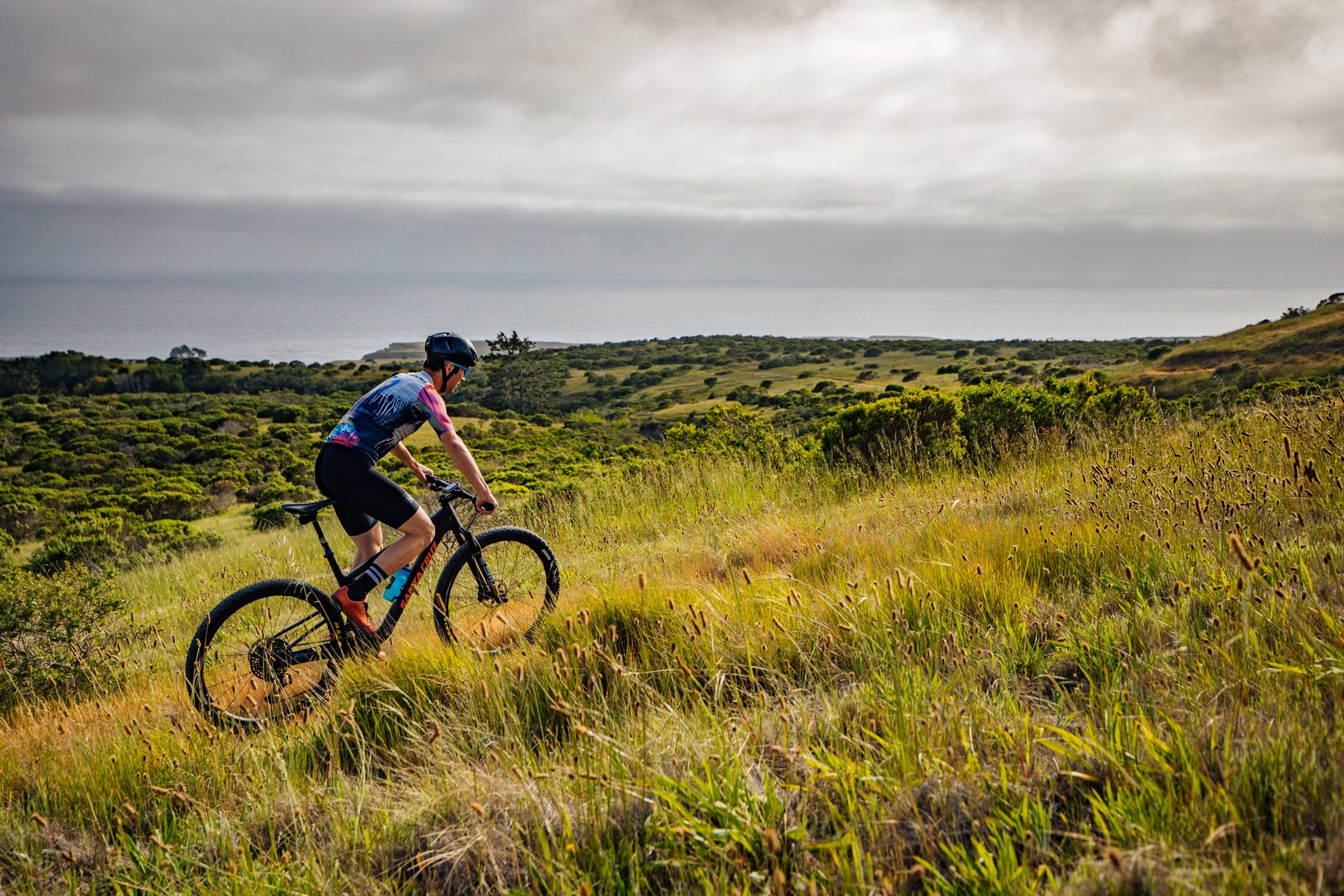 A cyclist rides a mountain bike through a grassy hillside with ocean views in the distance under a cloudy sky