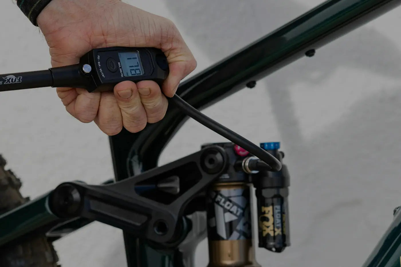 
A hand uses a digital shock pump to adjust the pressure on a FOX Float rear suspension, attached to a mountain bike frame