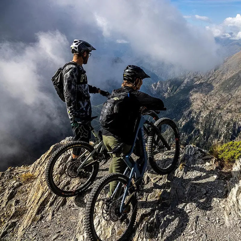 Two mountain bikers pause at a scenic cliff edge, admiring the rugged mountain landscape below